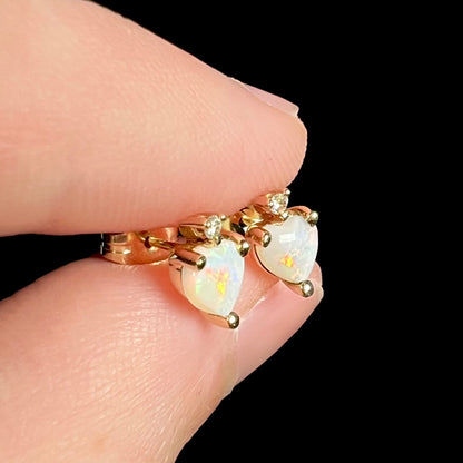 Angled close-up of 14k yellow gold opal heart stud earrings, held between fingers.  The opals have orange and red flashes.