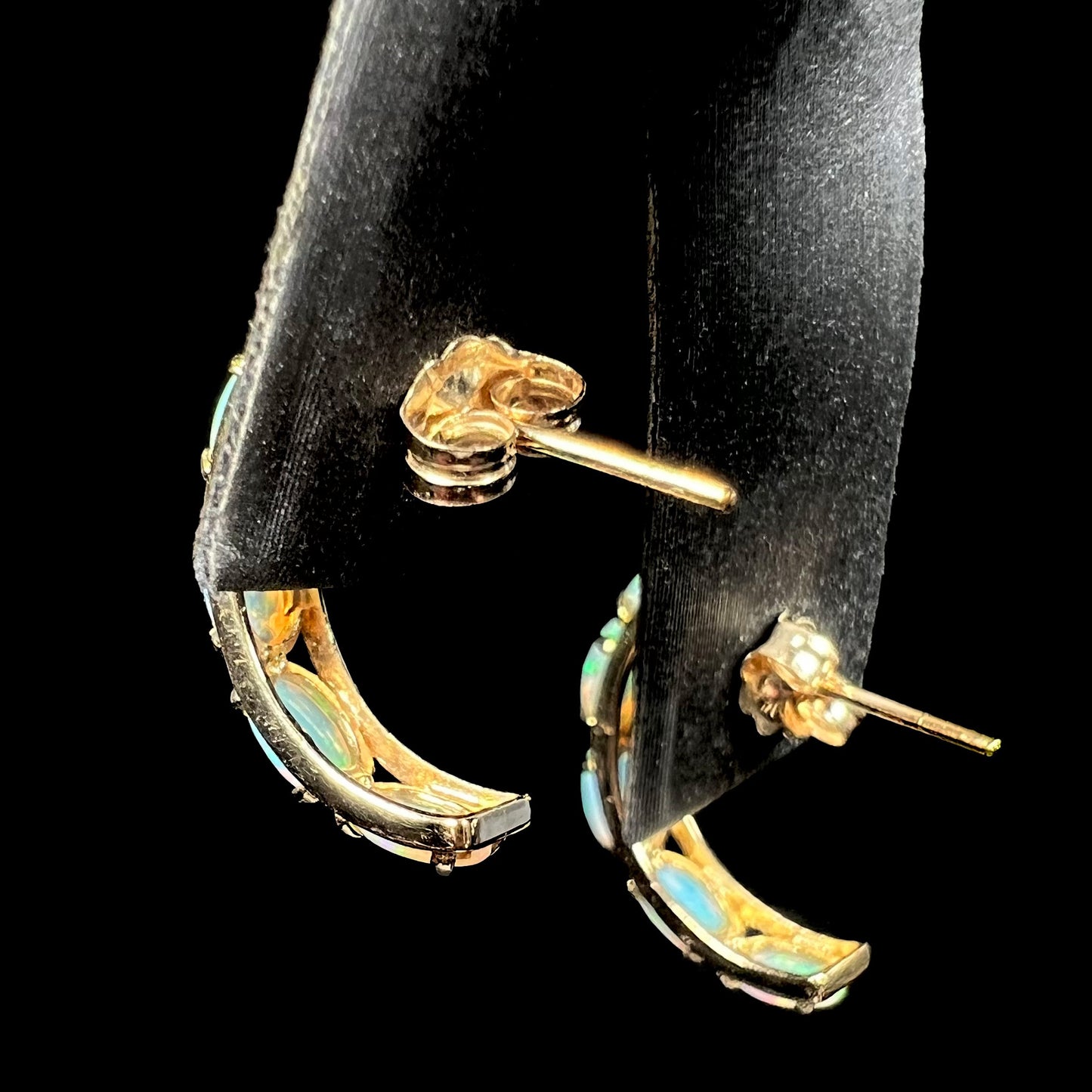 Back side of a pair of 14k yellow gold opal semi-hoop earrings focused on their push-back posts, shown on an earring display against a black background.