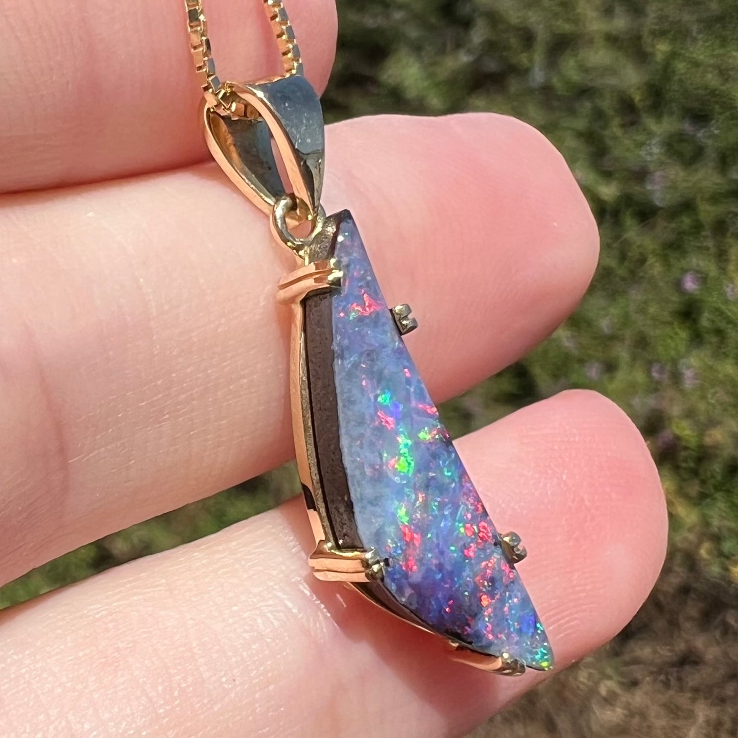 Angled view of 18k gold Australian boulder opal pendant in natural light, held in hand.