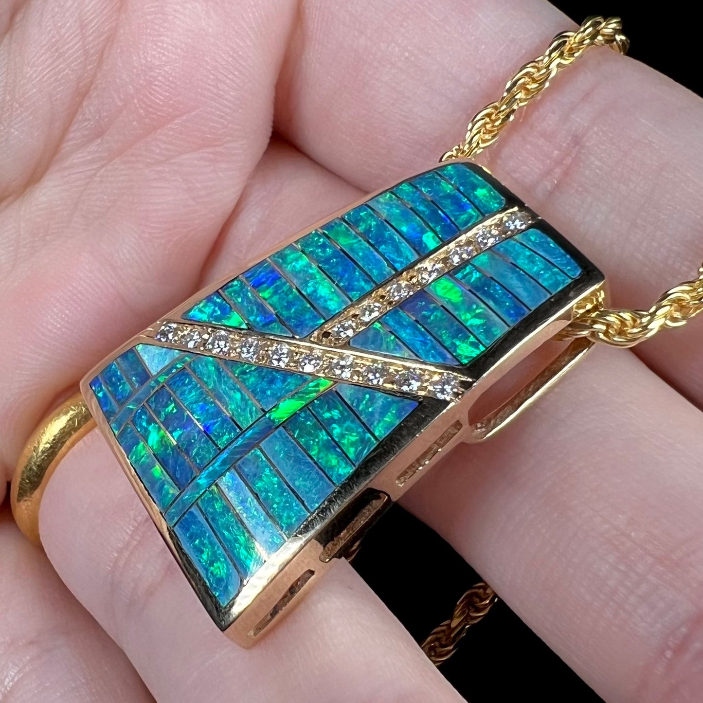An 18k yellow gold opal inlay pendant with diamond accents shown close-up from an angle held in a hand under white LED lighting.
