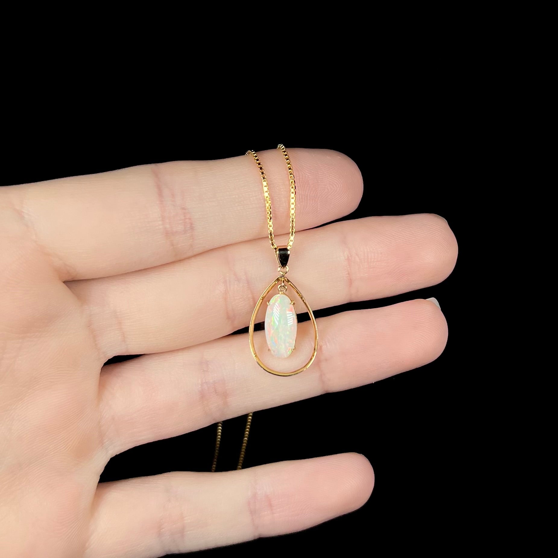 An 18k gold opal necklace resting on fingers against a black background, showing subtle red flashes in the opal.