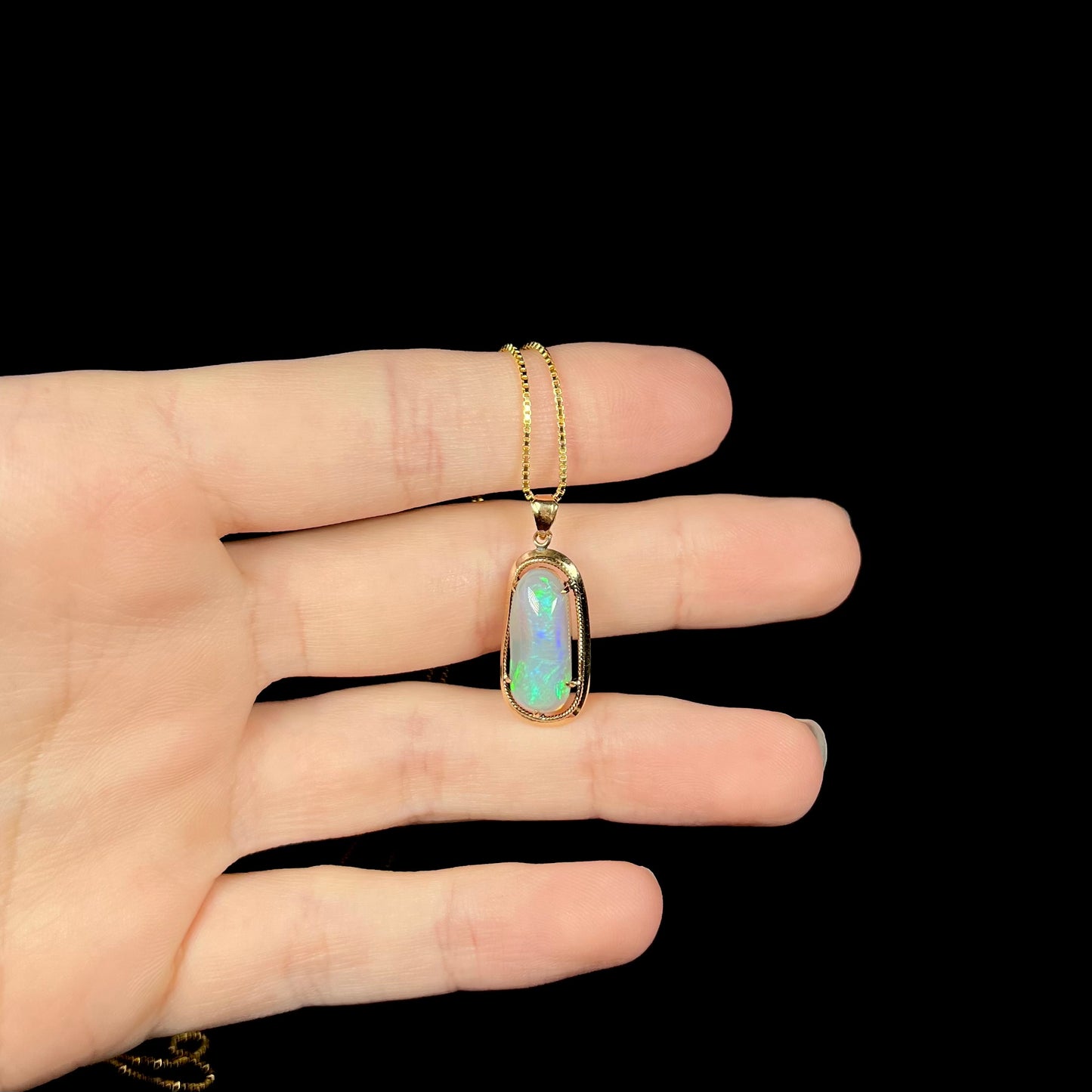 An 18k gold Lightning Ridge opal necklace resting on fingers against a black background, showing subtle blue and green flashes.