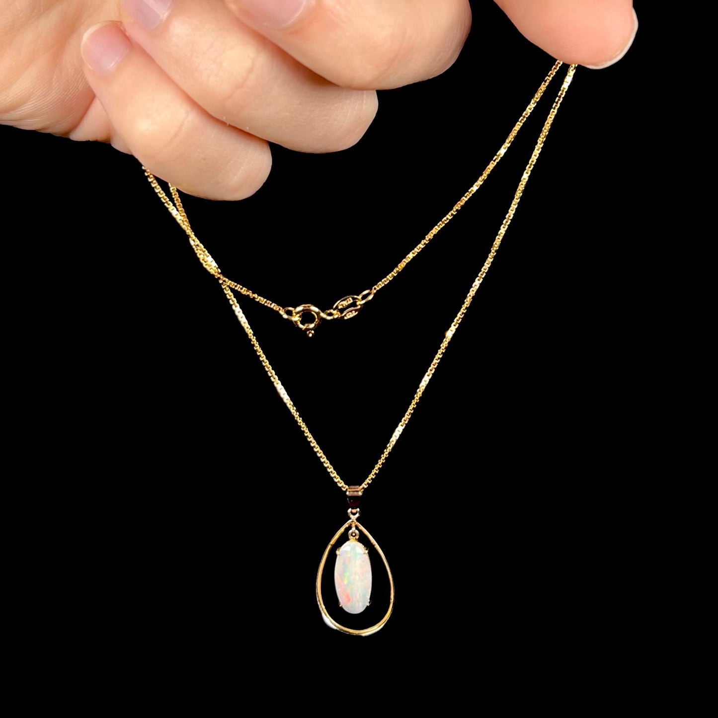 An 18k gold opal necklace being dangled in a hand against a black background, revealing subtle red flashes.