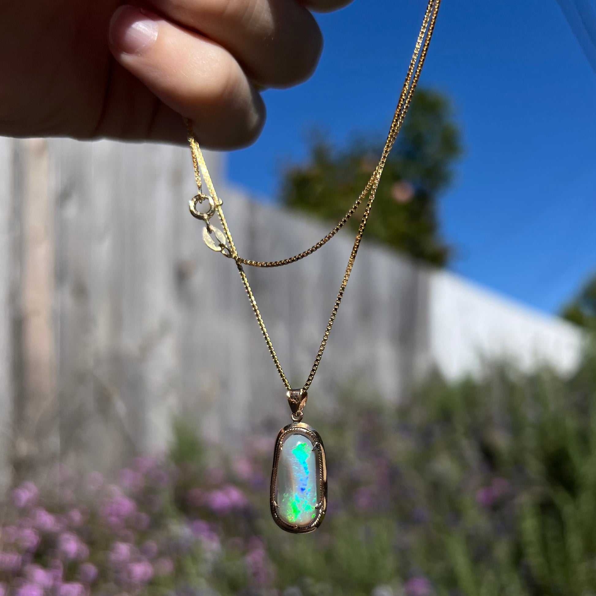 An 18k gold opal necklace outdoors in natural light, with bright green and blue colors appearing along the opal’s ridge.