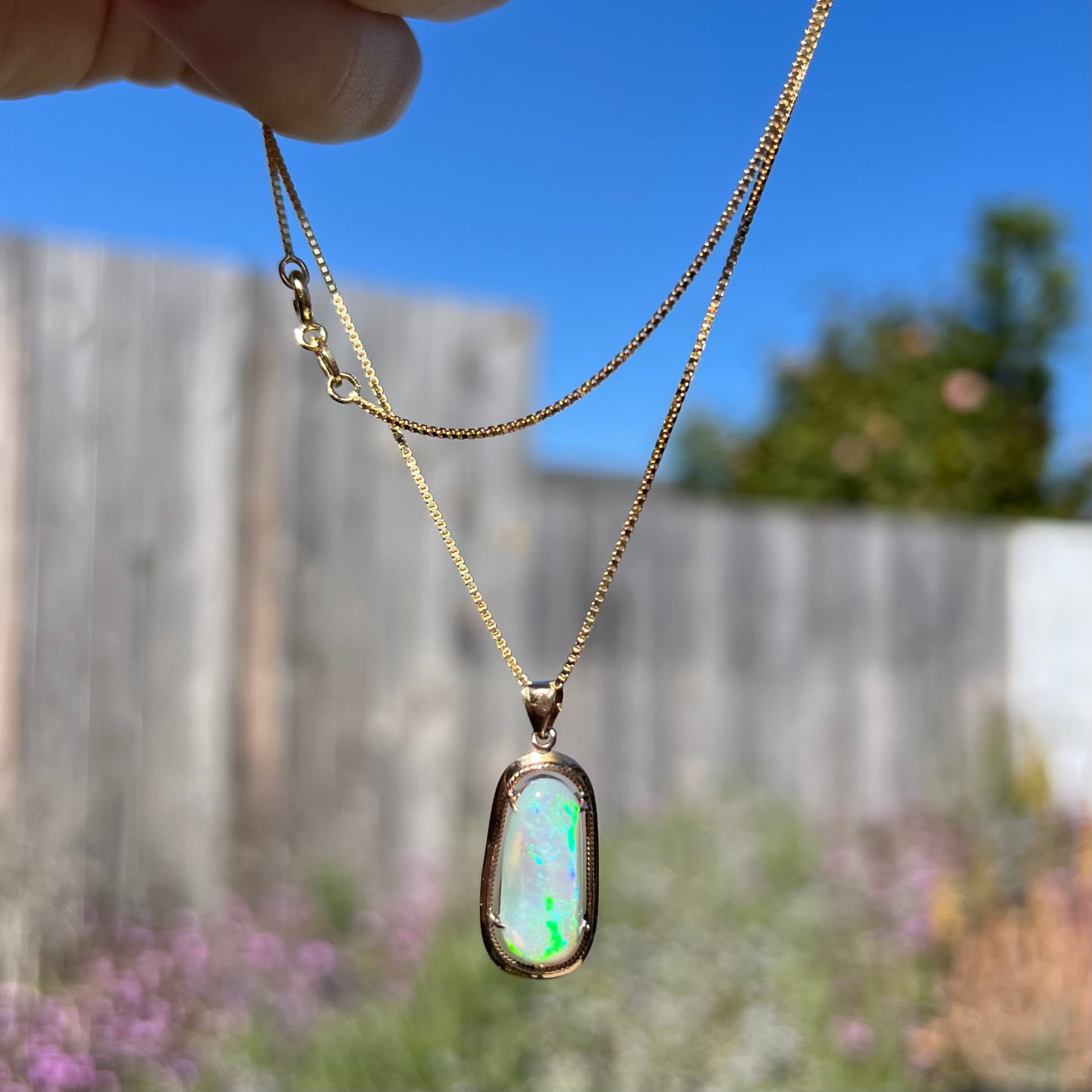 An 18k gold Lightning Ridge opal necklace being dangled outdoors in natural light, with overexposed flashes.