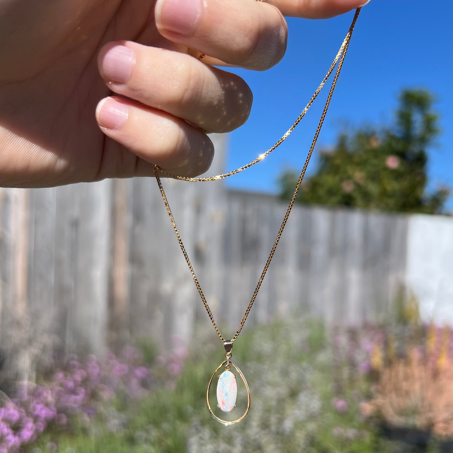 An 18k gold opal necklace being dangled outdoors in natural sunlight, showcasing red flashes in the opal.