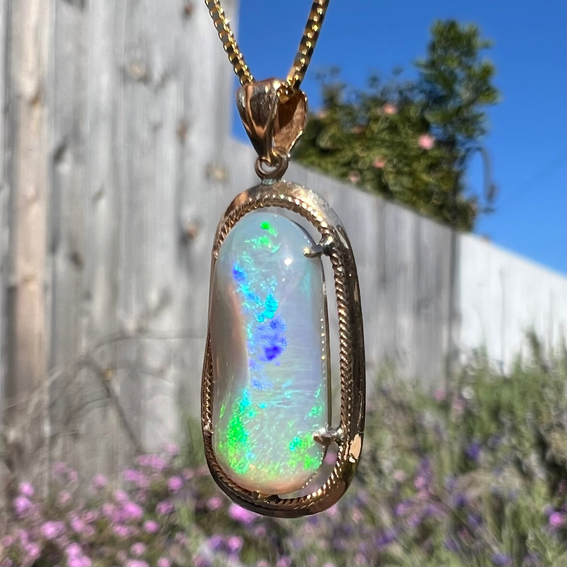 A macro view of an 18k gold Lightning Ridge opal necklace outdoors, twisted at an angle to highlight blue and green flashes.