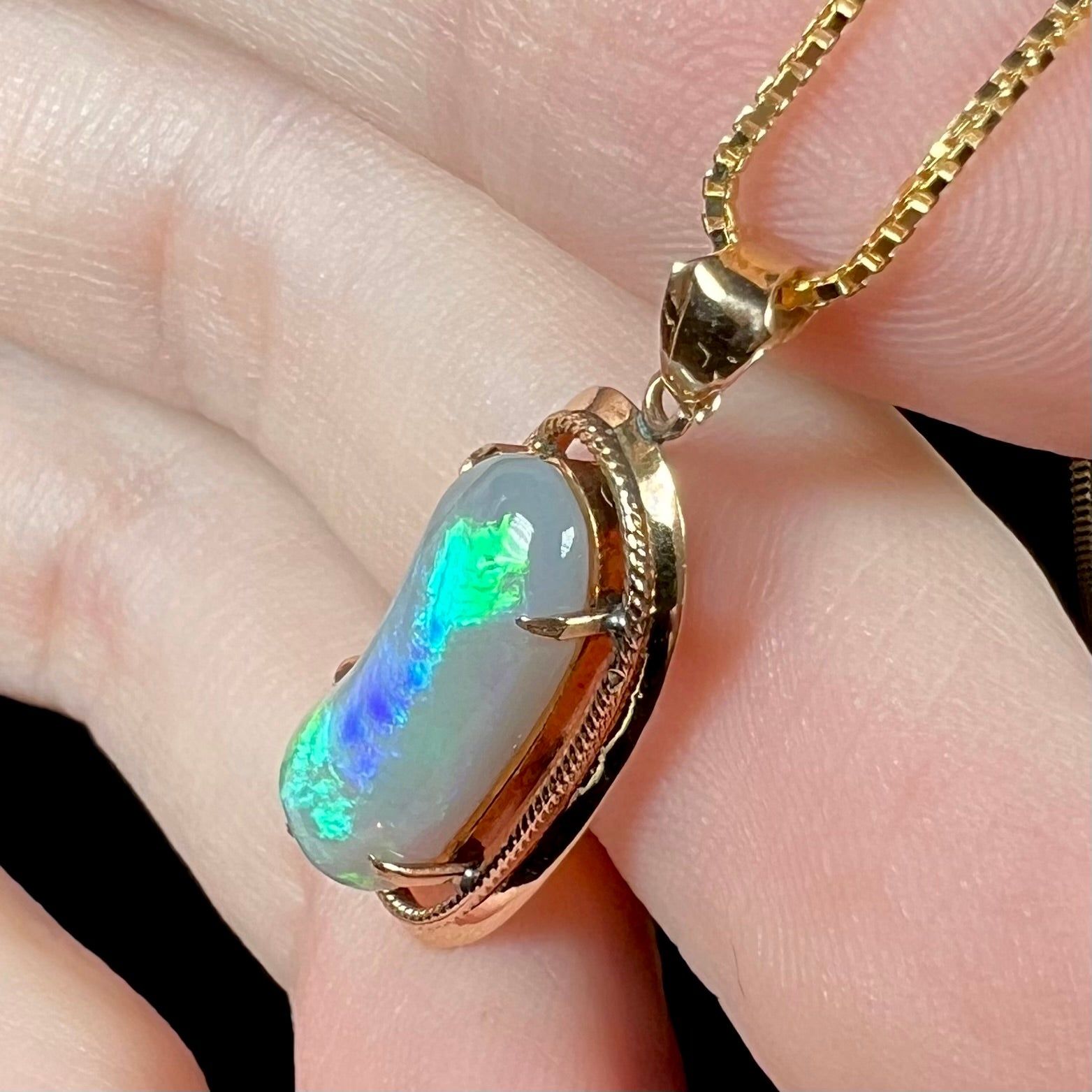 Slightly angled side view of an 18k gold Lightning Ridge opal necklace, showing a bean-like shape with blue and green flashes running down its ridge.