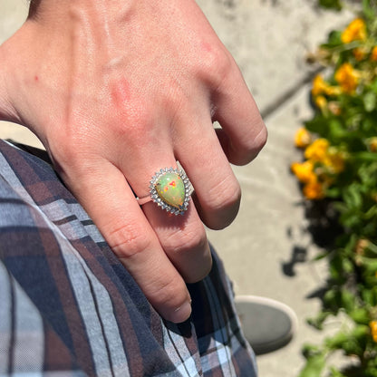 Hand resting on person's hip in natural light while wearing a pear shaped 18k white gold Ethiopian opal and diamond halo ring.
