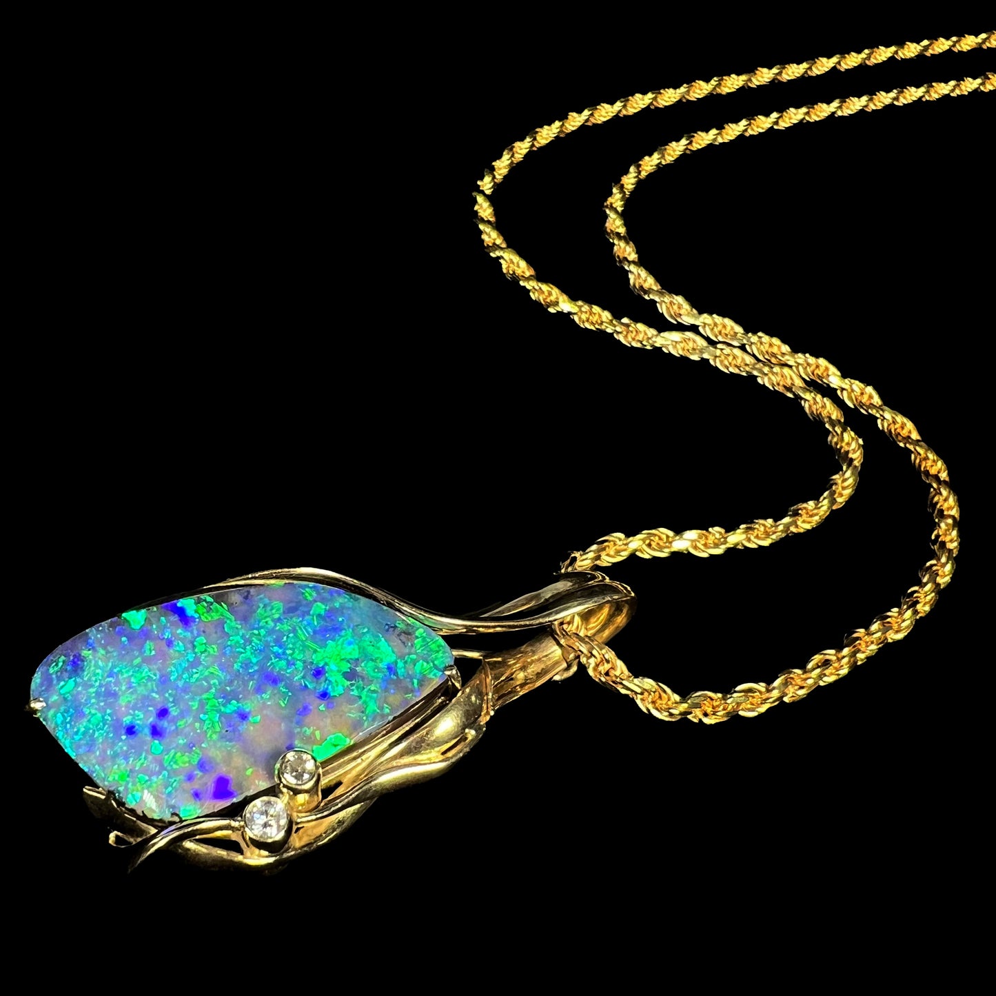 Angled close-up of 18k yellow gold boulder opal and diamond necklace with blue and green play of color laying on black background.