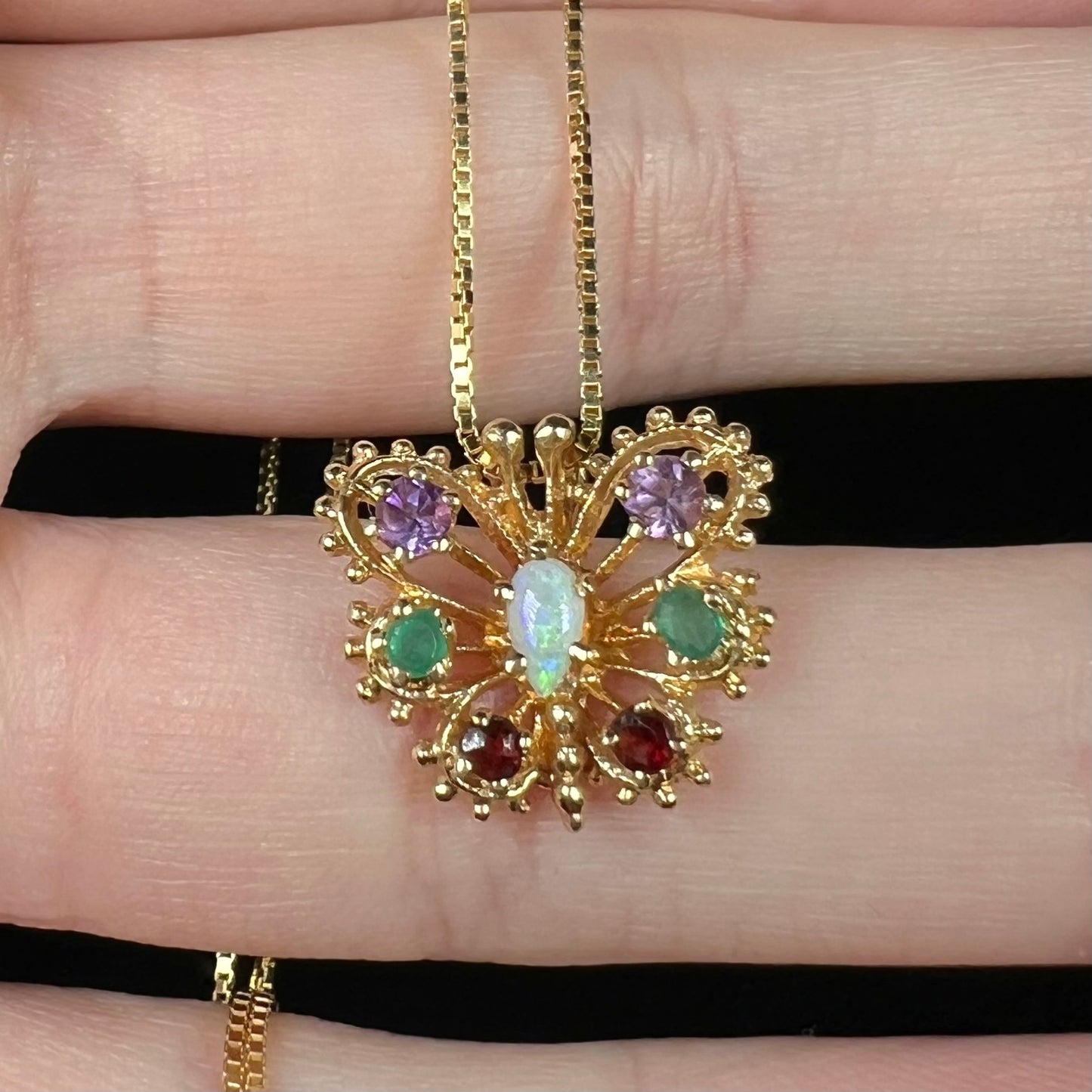 Close-up of 1960s era yellow gold and multi-gemstone butterfly pendant held in hand, shown with box-style chain.