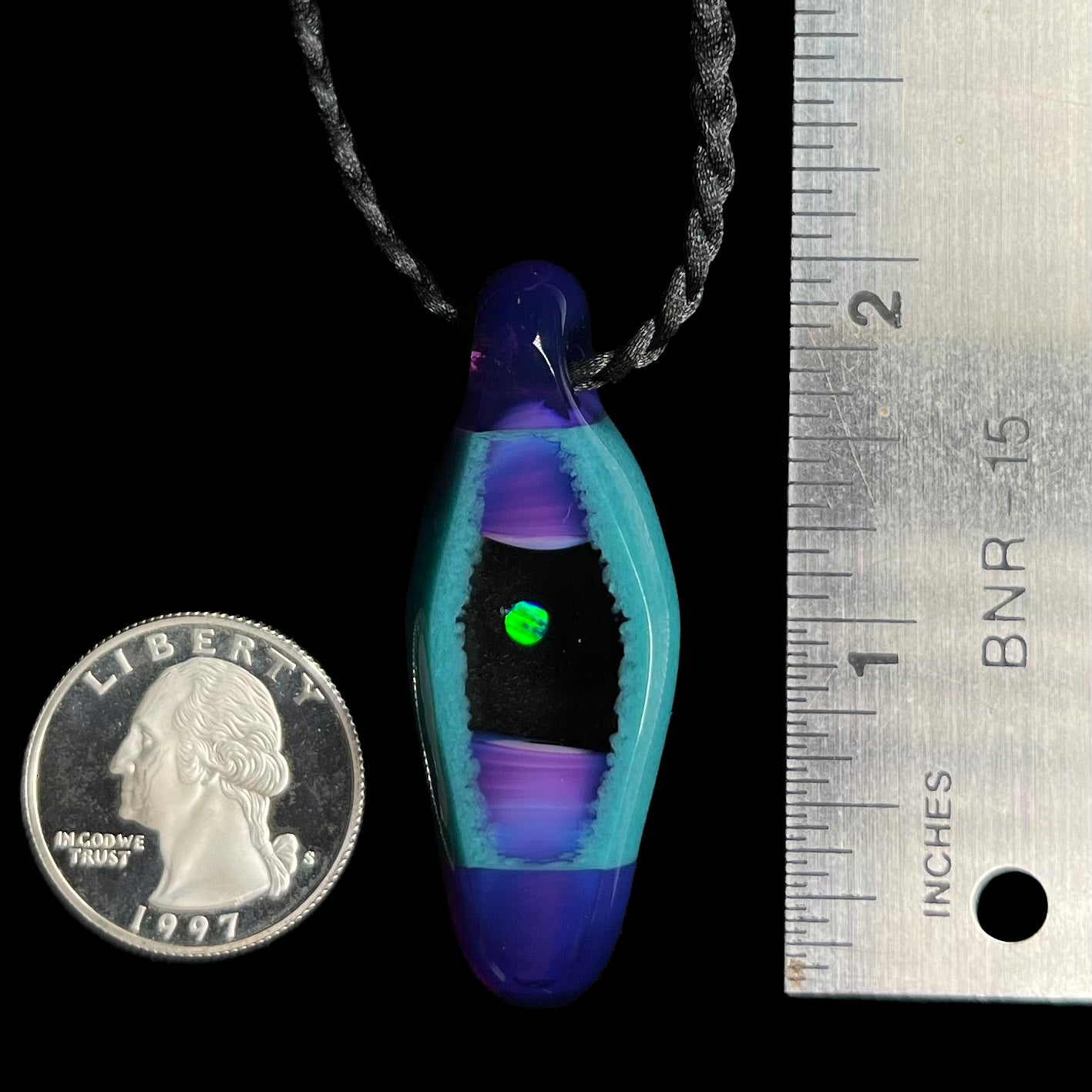 A 2 inch-long opal in glass pendant displayed next to a quarter coin and ruler for scale.