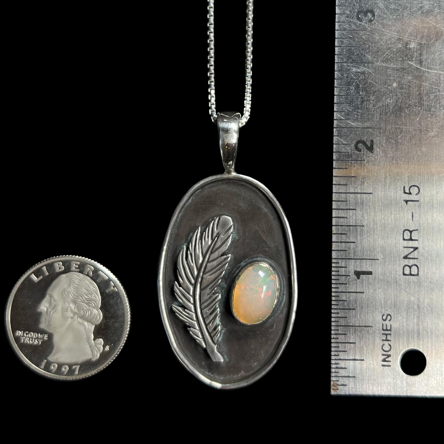 A 2 inch-long sterling silver men's opal and feather design pendant shown on a black background next to a ruler and US quarter coin.