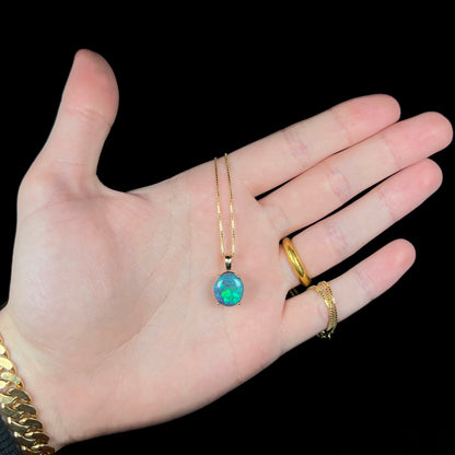 Hand holding 3.80ct oval shaped Australian black opal solitaire pendant mounted in 14k gold; a gold-tone chain wraps around hand's pinky finger