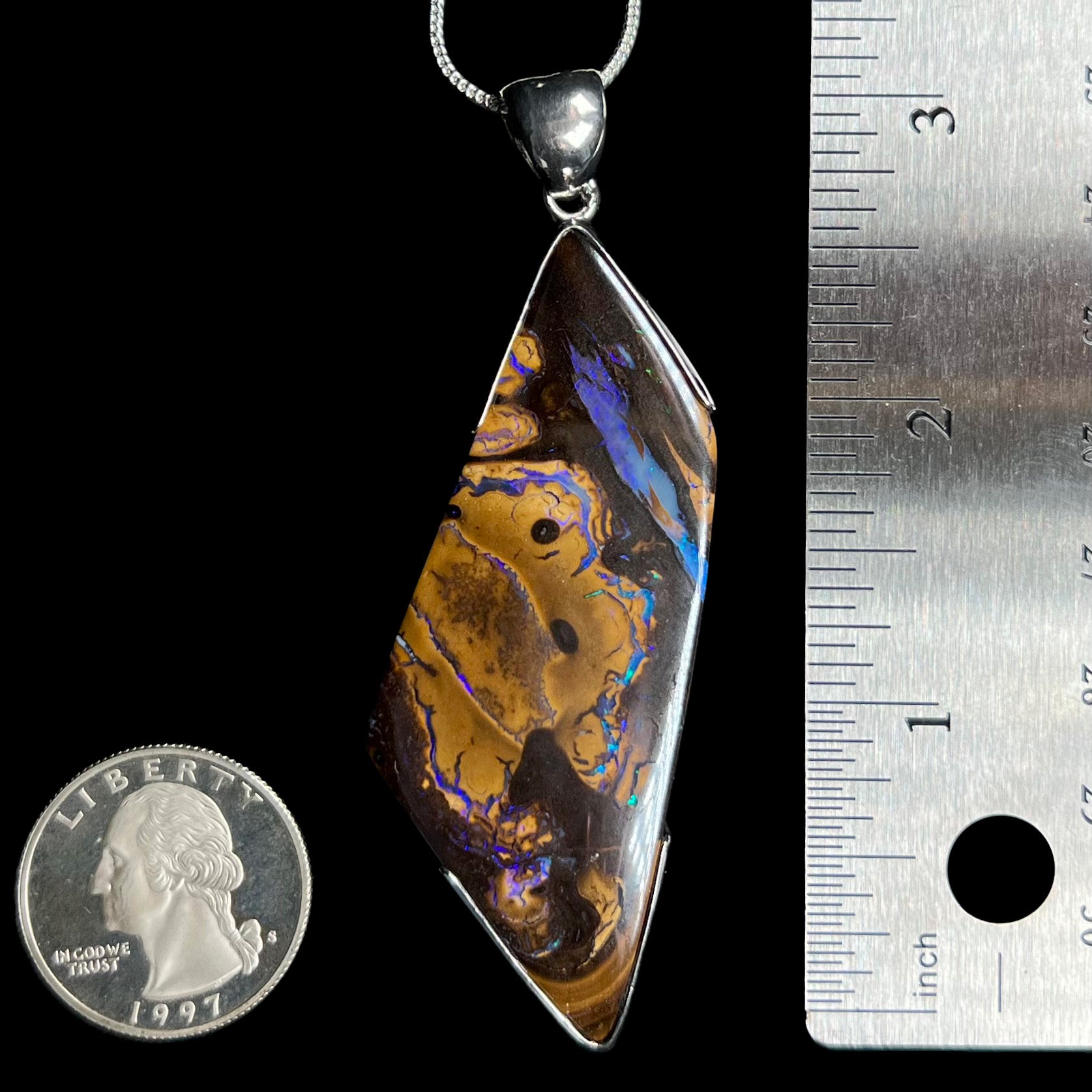 A large Australian boulder opal pendant in silver measuring over 3 inches long next to US quarter coin and ruler