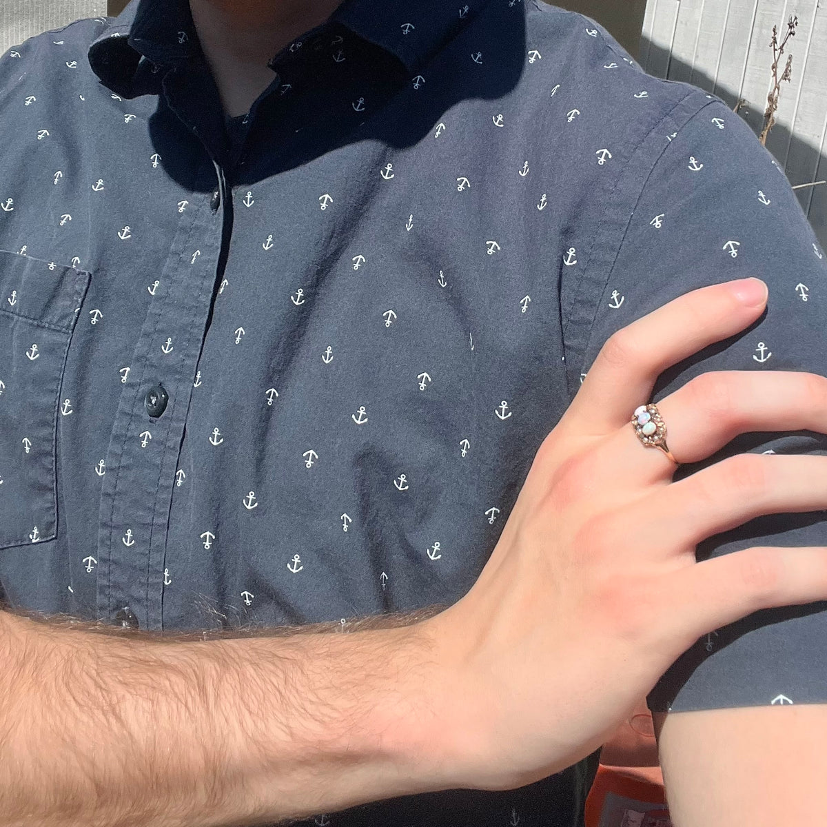 Close-up of person resting hand on opposing shoulder in sunlight wearing a Victorian three-stone opal and seed pearl halo ring in 9k gold
