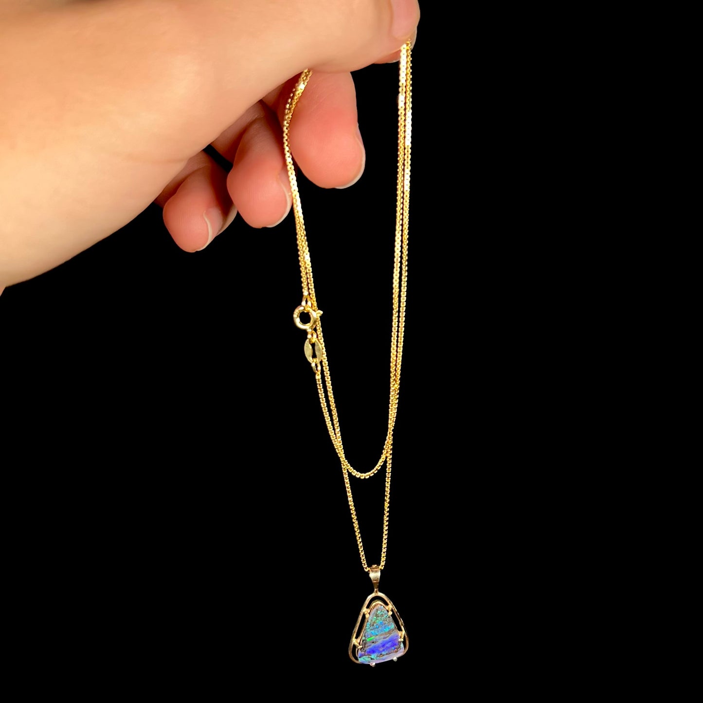 A dainty Duck Creek opalized wood necklace in 9k gold, shown dangled from a hand by its chain.