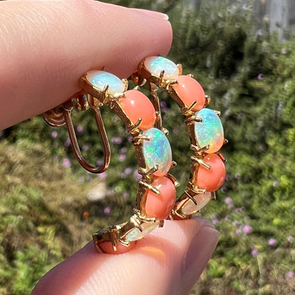 Side view of 14k gold semi-hoop earrings with alternating prong-set Australian crystal opals and pink coral cabochons, held between two fingers in sunlight