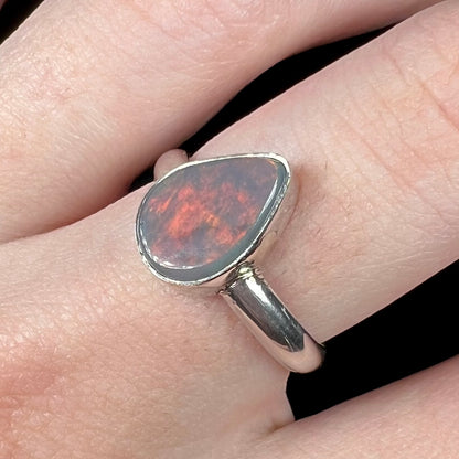 Close-up of an American-made sterling silver Australian dark opal solitaire ring, worn on a finger.  A broad flash of soft red color is seen.