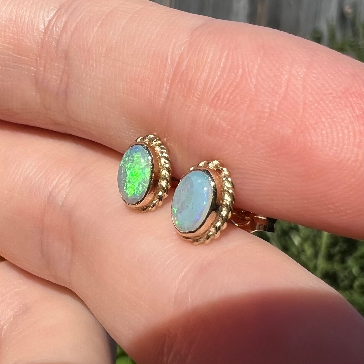 Pair of Australian opal stud earrings with green flash of color made by American company, Carla Corp., shown from angle in sunlight.