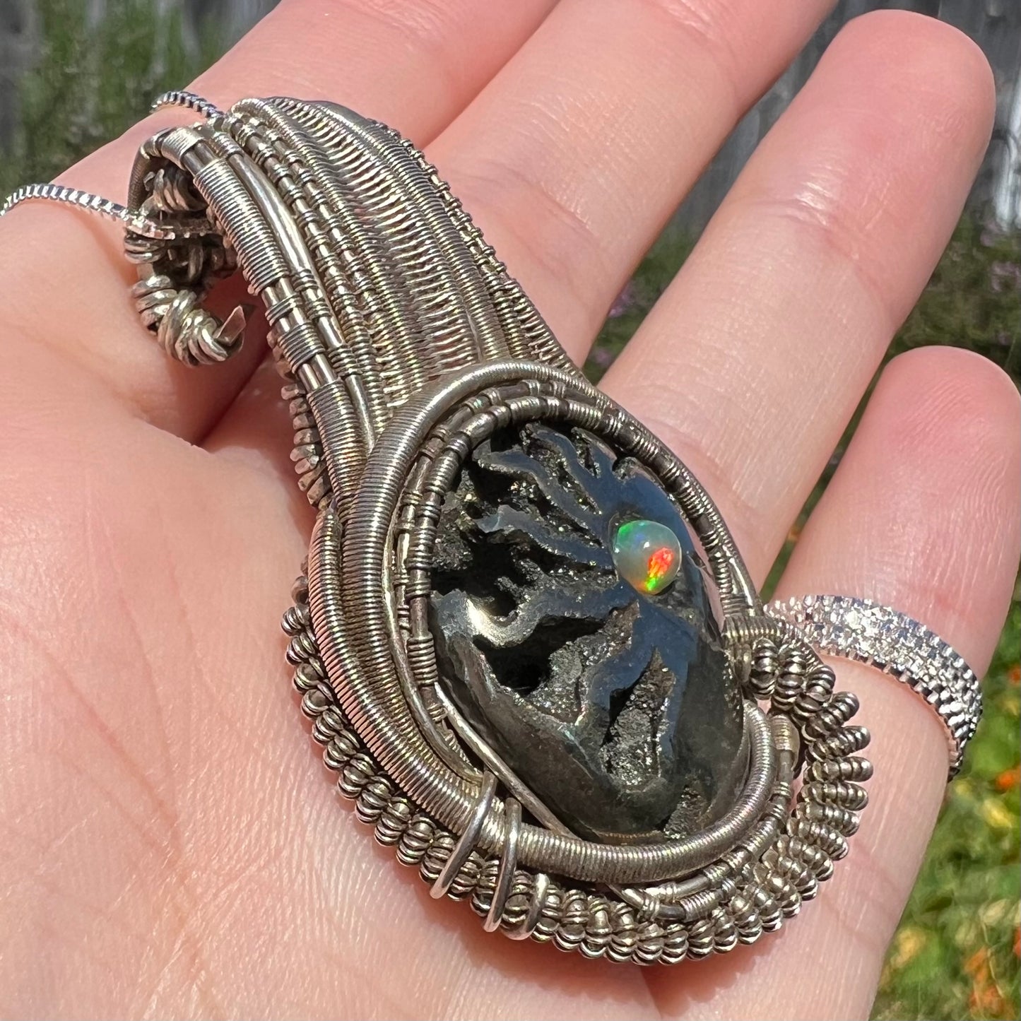 Angled close-up of silver wire wrapped ammonite shell pendant set with opal accent, held in hand.  The opal shows a red flash in sunlight.