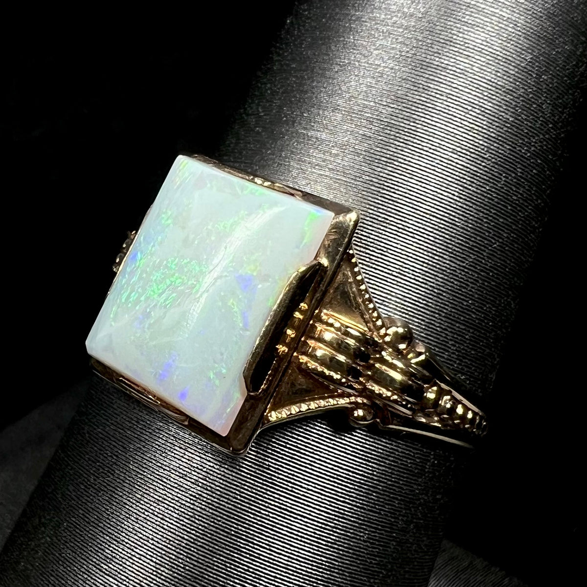 Angled close-up of an antique c.1920s era Art Deco opal solitaire ring in 10k gold on a ring display, focused on ring's ornate milgrain-textured shoulder
