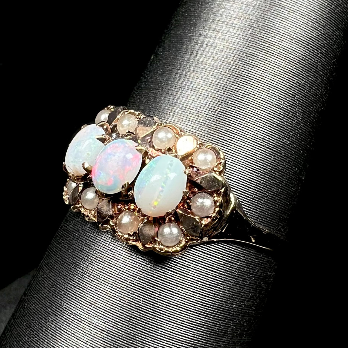 Angled close-up of antique 9k gold three-stone opal ring with halo of seed pearl accents, shown on ring display