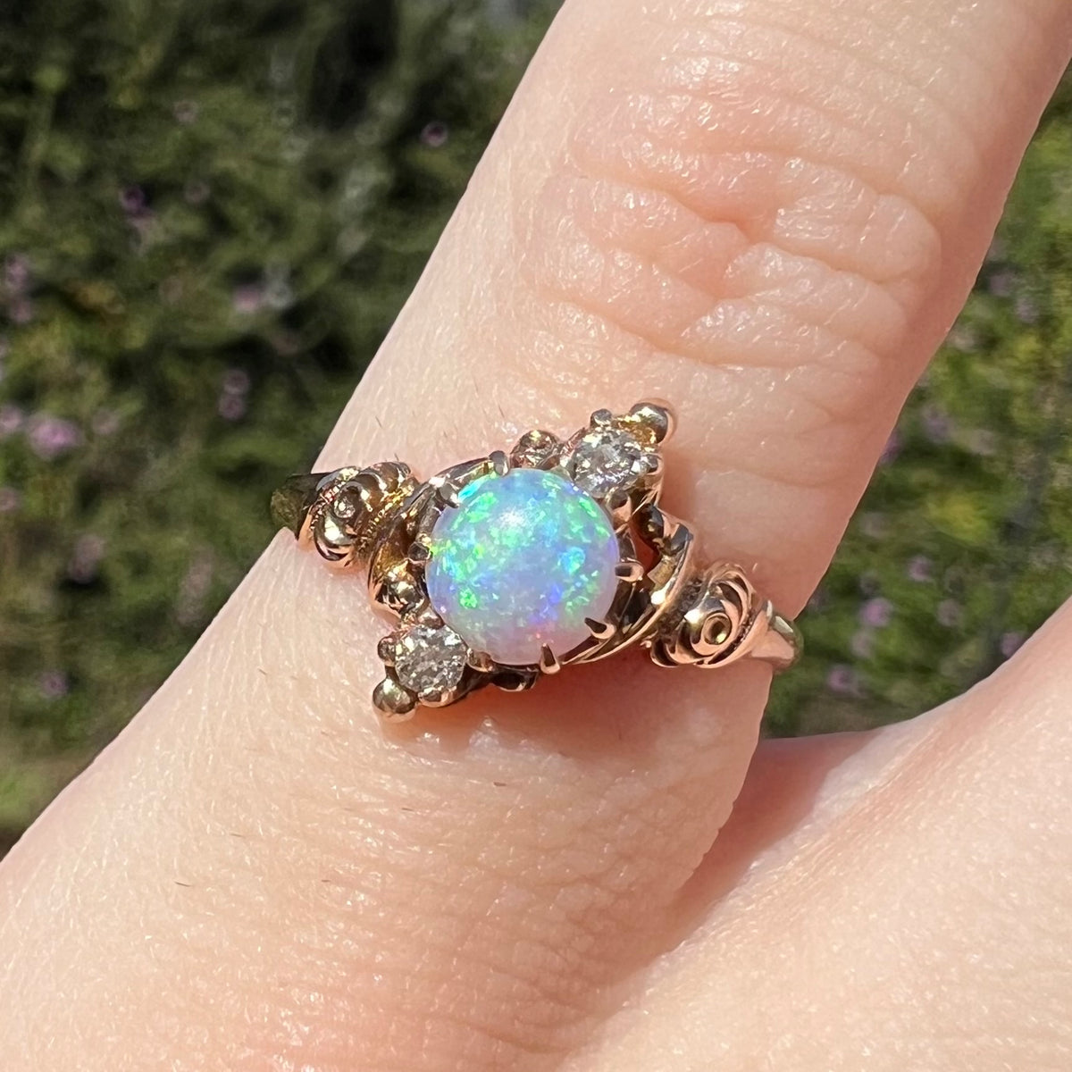 Close-up of Art Nouveau era opal and diamond pinky ring, worn on finger in sunlight; the opal shows green and blue colors
