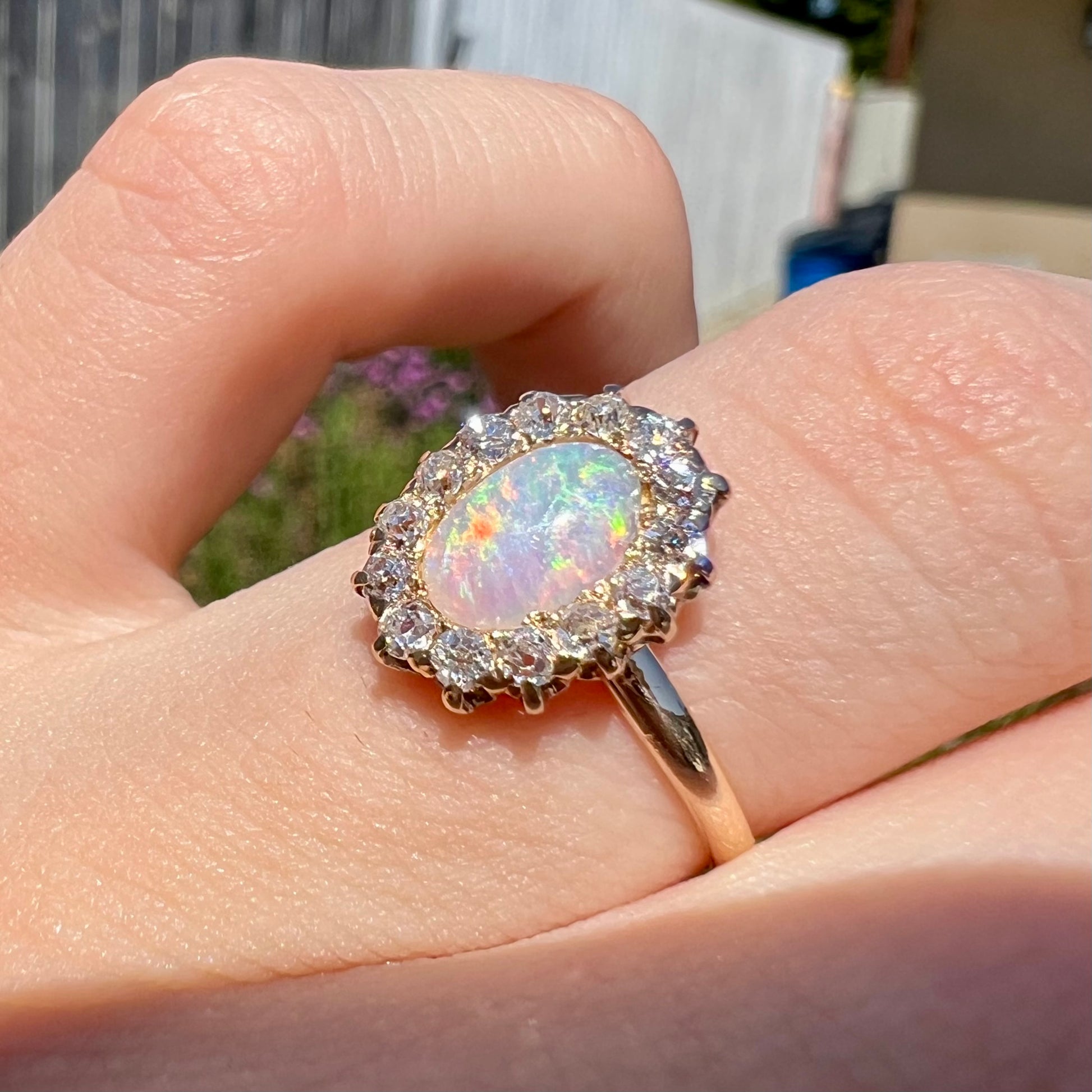 An antique opal and diamond halo engagement ring being worn on a hand in the sunlight.  The ring is yellow gold.