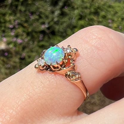 Green colors visible across surface of round opal mounted in antique 10k gold pinky ring, worn on finger and shown from angle in sunlight; the ring has a rose design motif on its shoulder