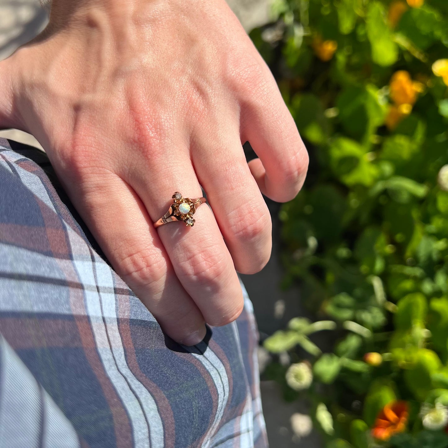 Hand wearing an antique Victorian opal and diamond three-stone ring while resting on hip in sunlight against a leafy green backdrop.