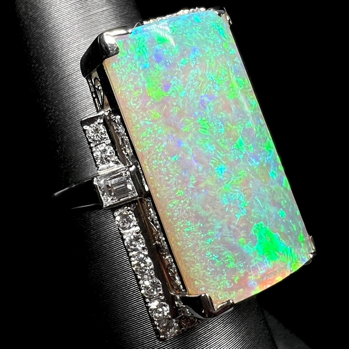 An Art Deco-inspired portrait-cut Australian light opal ring shown on ring display from angle, a partial platinum frame set with round and emerald cut diamonds in view.