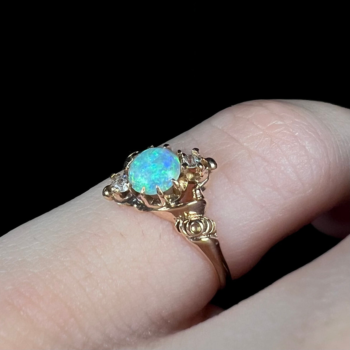 Angled close-up of Art Nouveau style opal and diamond pinky ring in 10k gold with rose design shoulders, worn on pinky finger