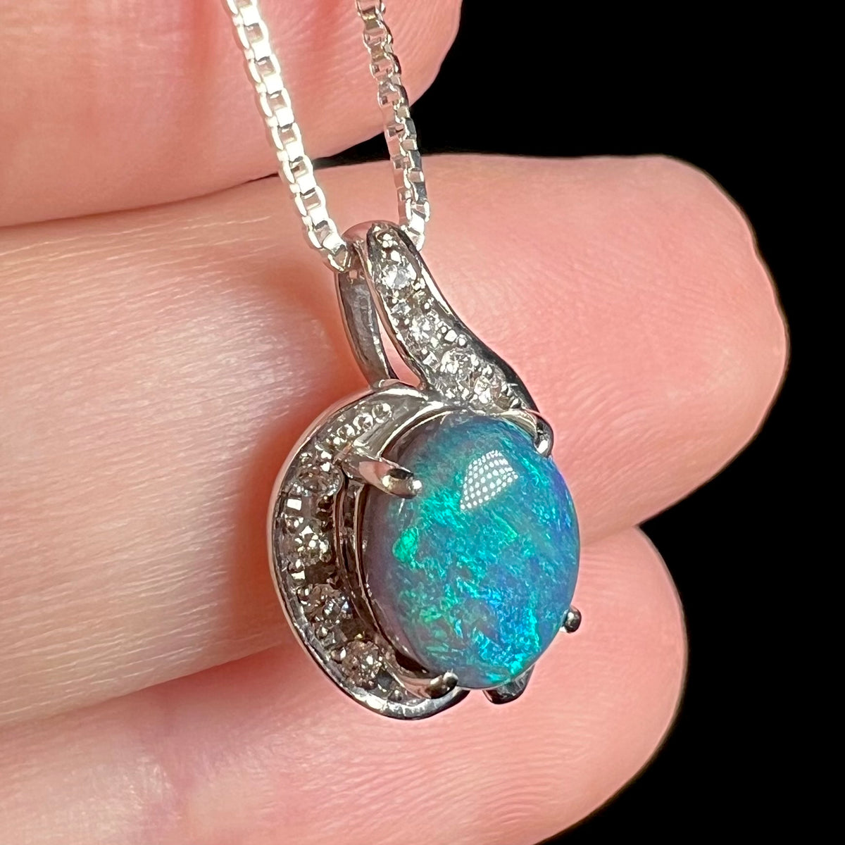 Angled close-up of an Australian black semi-crystal opal pendant with diamond accents, held in a hand.  The opal has blue and green play-of-color.