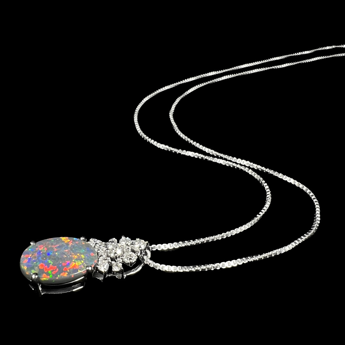 A platinum Australian black opal and diamond necklace with red flashes of play-of-color shown laying on black background, its chain flowing into the distance.