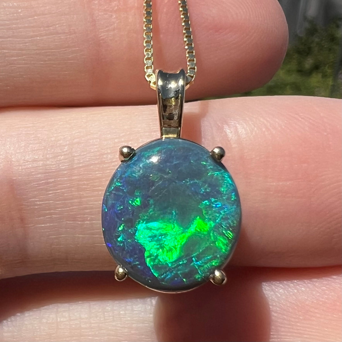 Close-up of oval shaped Australian black opal pendant in 14k gold, resting on fingers in natural sunlight; the opal has bright green flashes of color