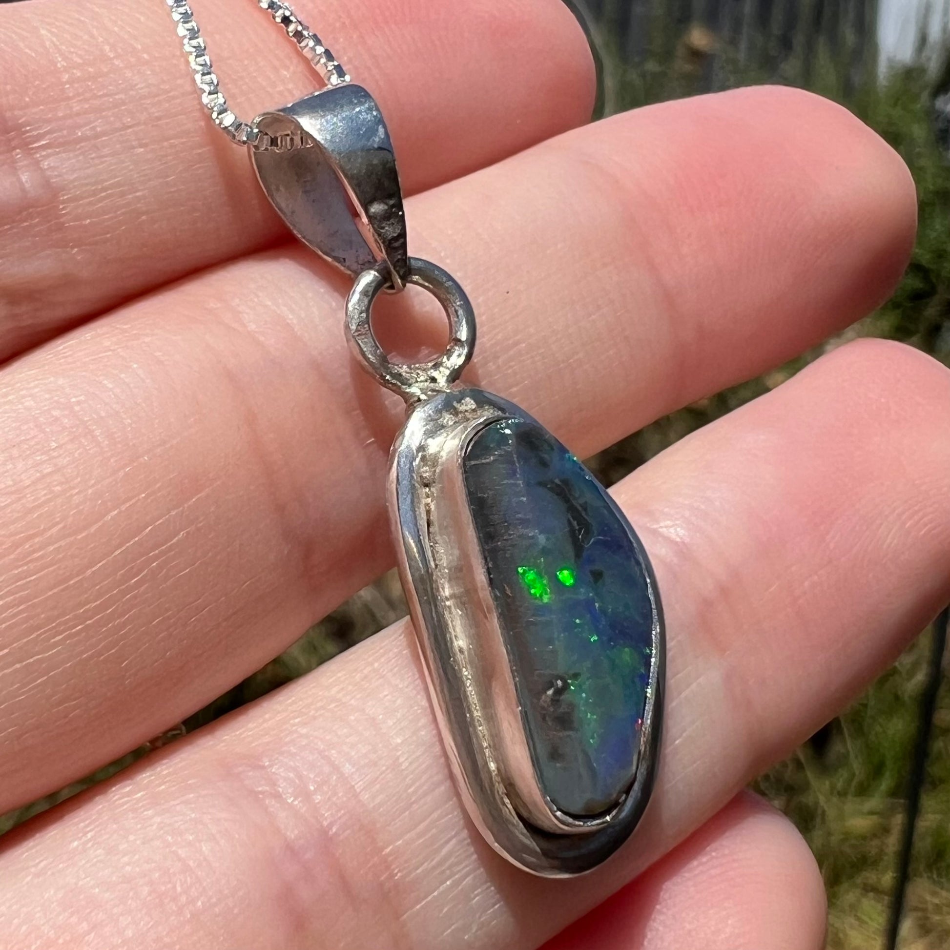 Angled close-up of a sterling silver Australian black opal pendant held in natural sunlight, green flashes of color visible in the opal.