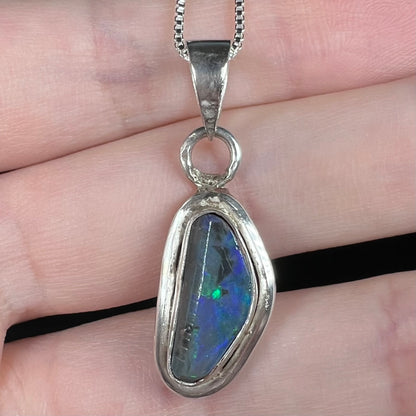 Close-up of a sterling silver Australian black opal pendant held on fingers; the opal has one green flash of color.