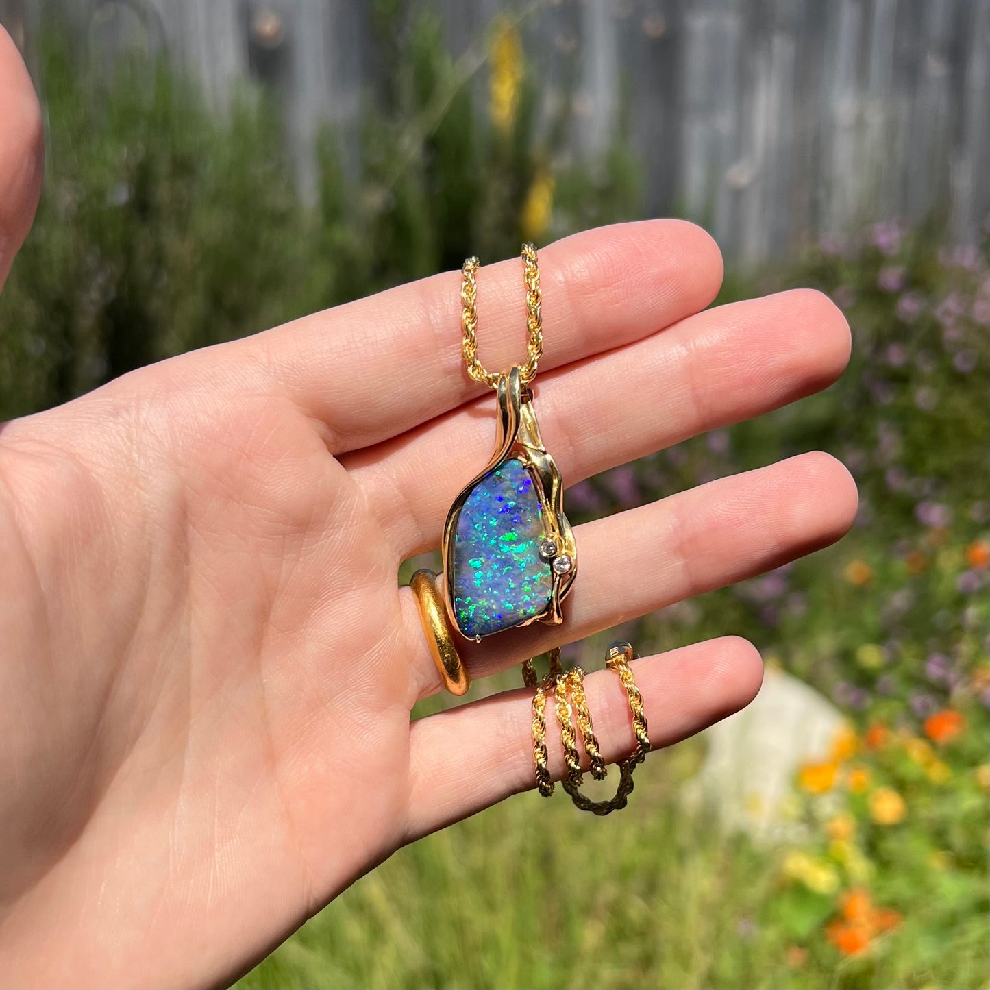 Hand holding a large Australian boulder opal and diamond statement pendant in natural light. The opal has green and blue flashes of color.