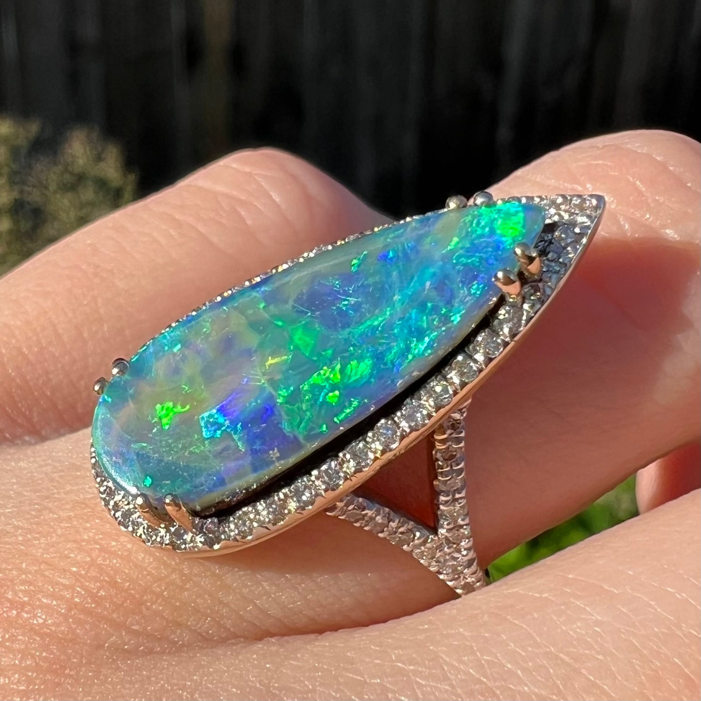 Angled close-up of a pear-shaped Australian boulder opal ring with a split shank design, worn on a finger in sunlight.