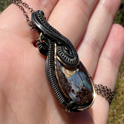 Angled close-up of an Australian boulder opal pendant featuring a handmade two-tone blackened sterling silver wire wrapped design, held in a hand in direct sunlight.