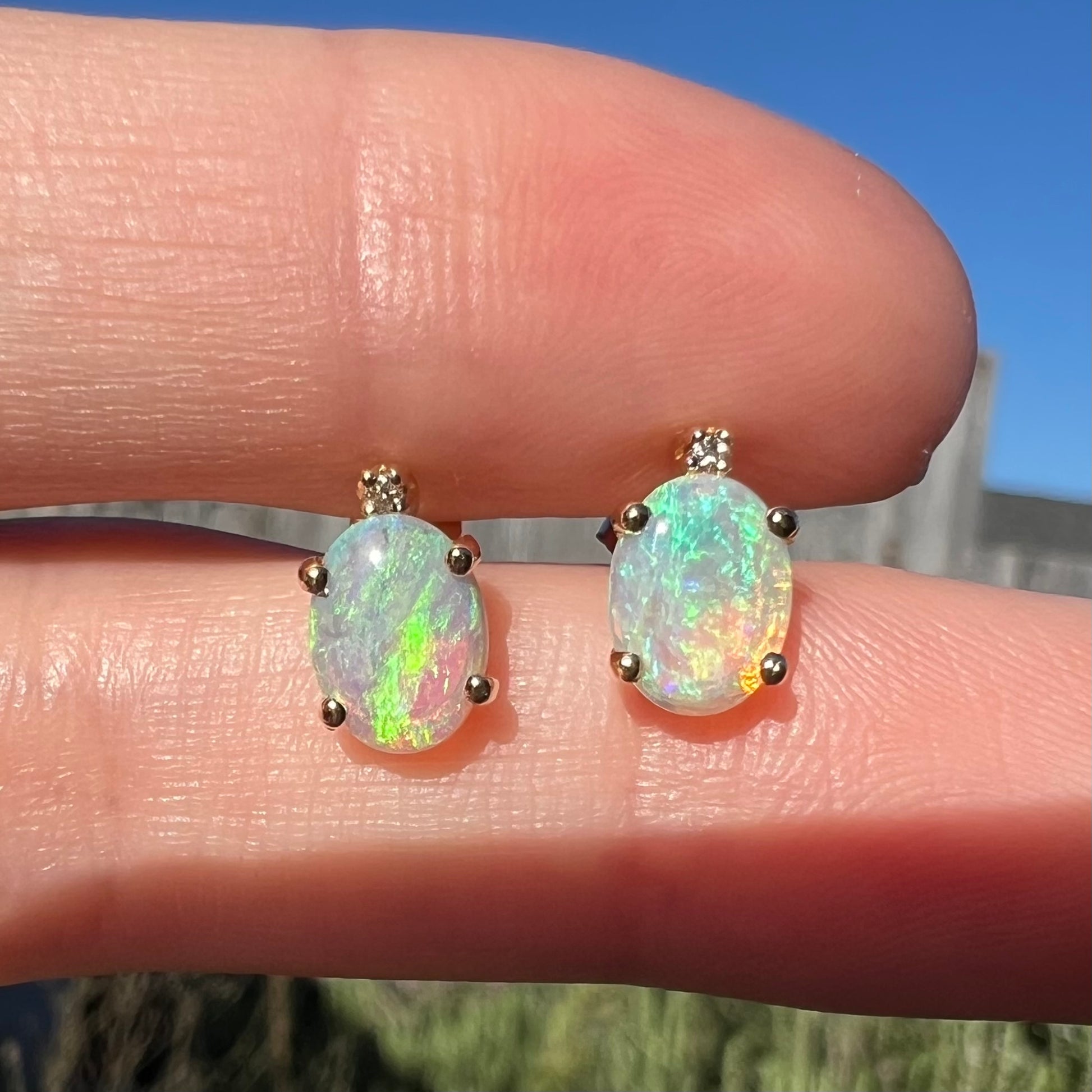 A pair of Australian crystal opal and diamond stud earrings in gold with green color play shown close-up in the sunlight.