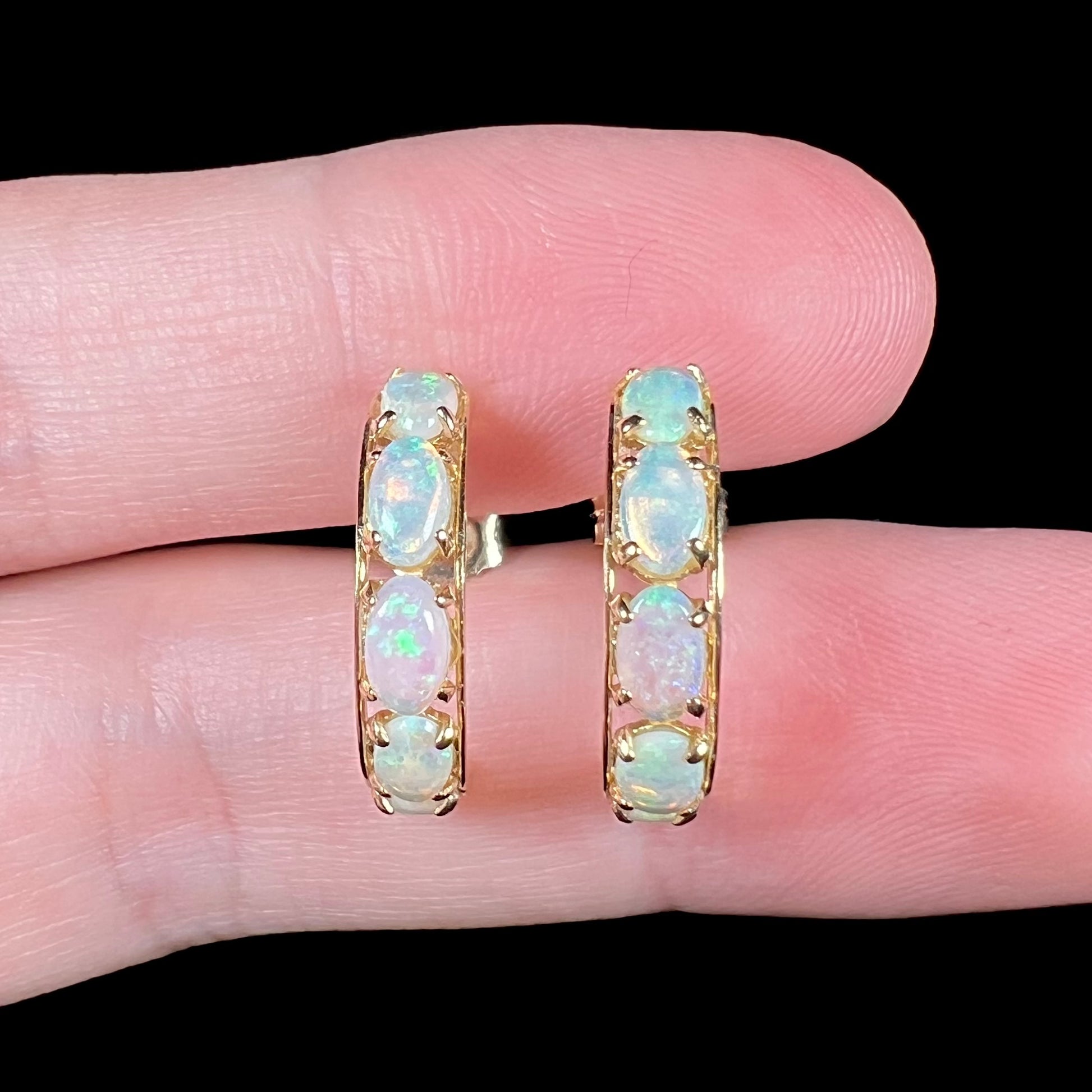 Close-up of yellow gold Australian crystal opal hoop earrings held between two fingers under studio LED lighting against a black backdrop.