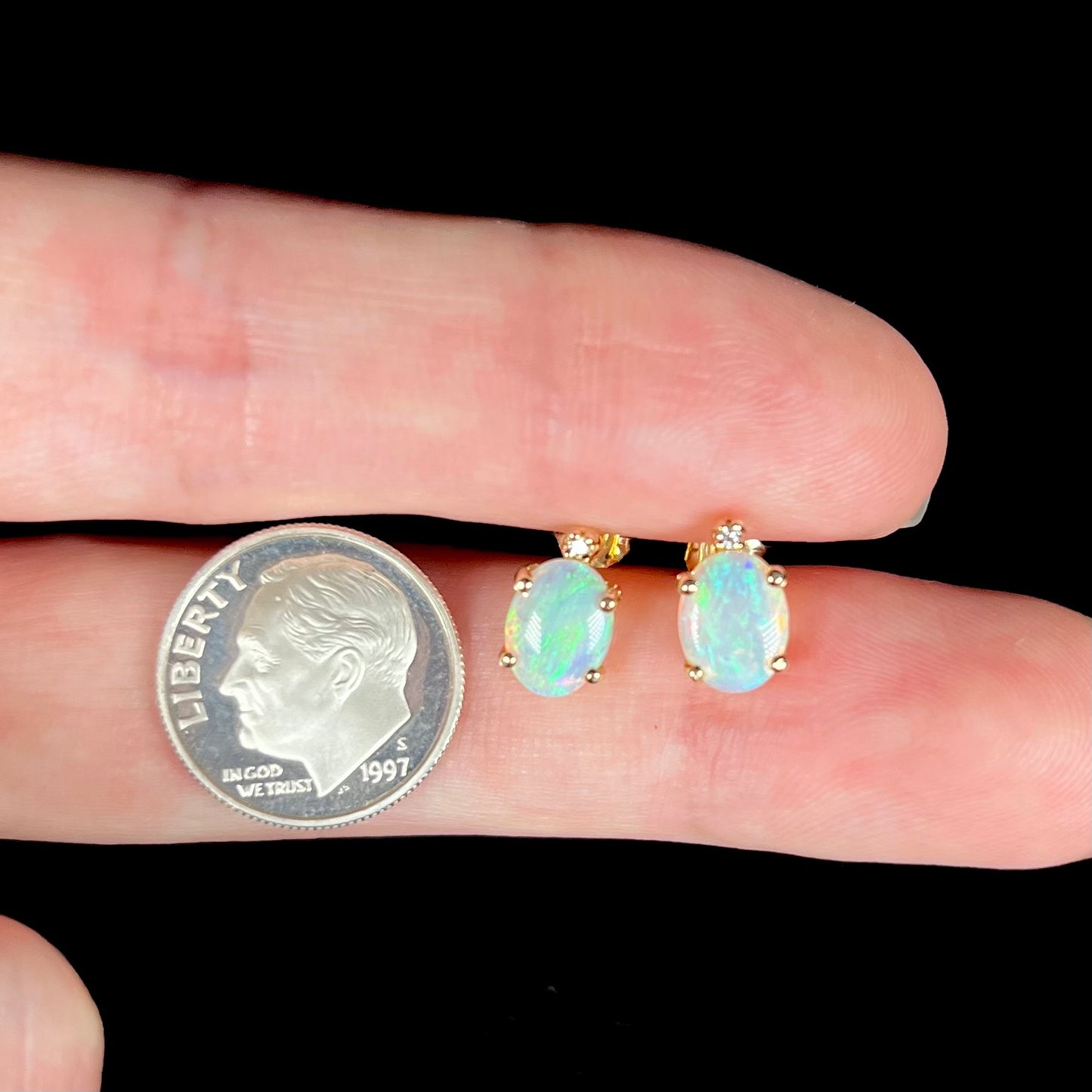 A pair of Australian crystal opal and diamond stud earrings in gold shown between two fingers next to a dime coin.