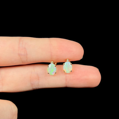 A pair of Australian crystal opal and diamond stud earrings in gold with green-blue color play shown held between two fingers against a black background.