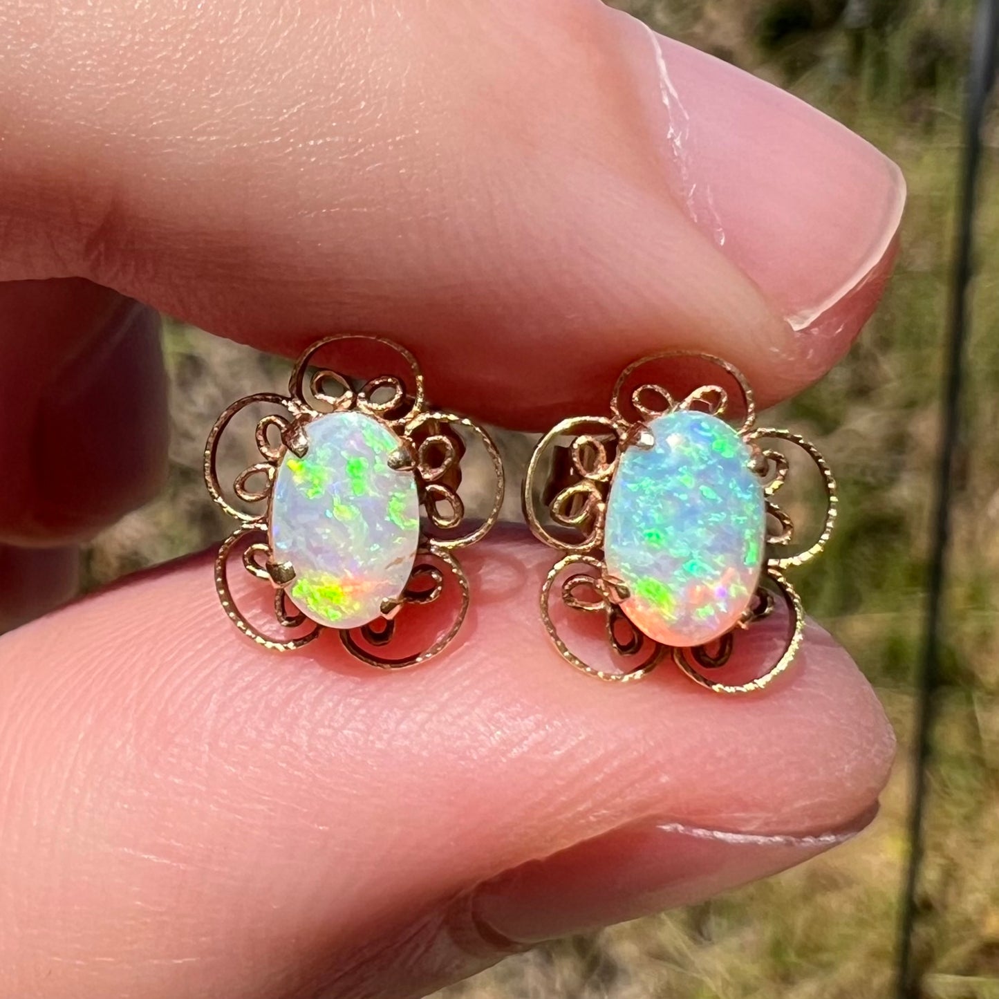 Close-up of Australian crystal opal stud earrings in 14k gold with flower design, held between fingers in sunlight