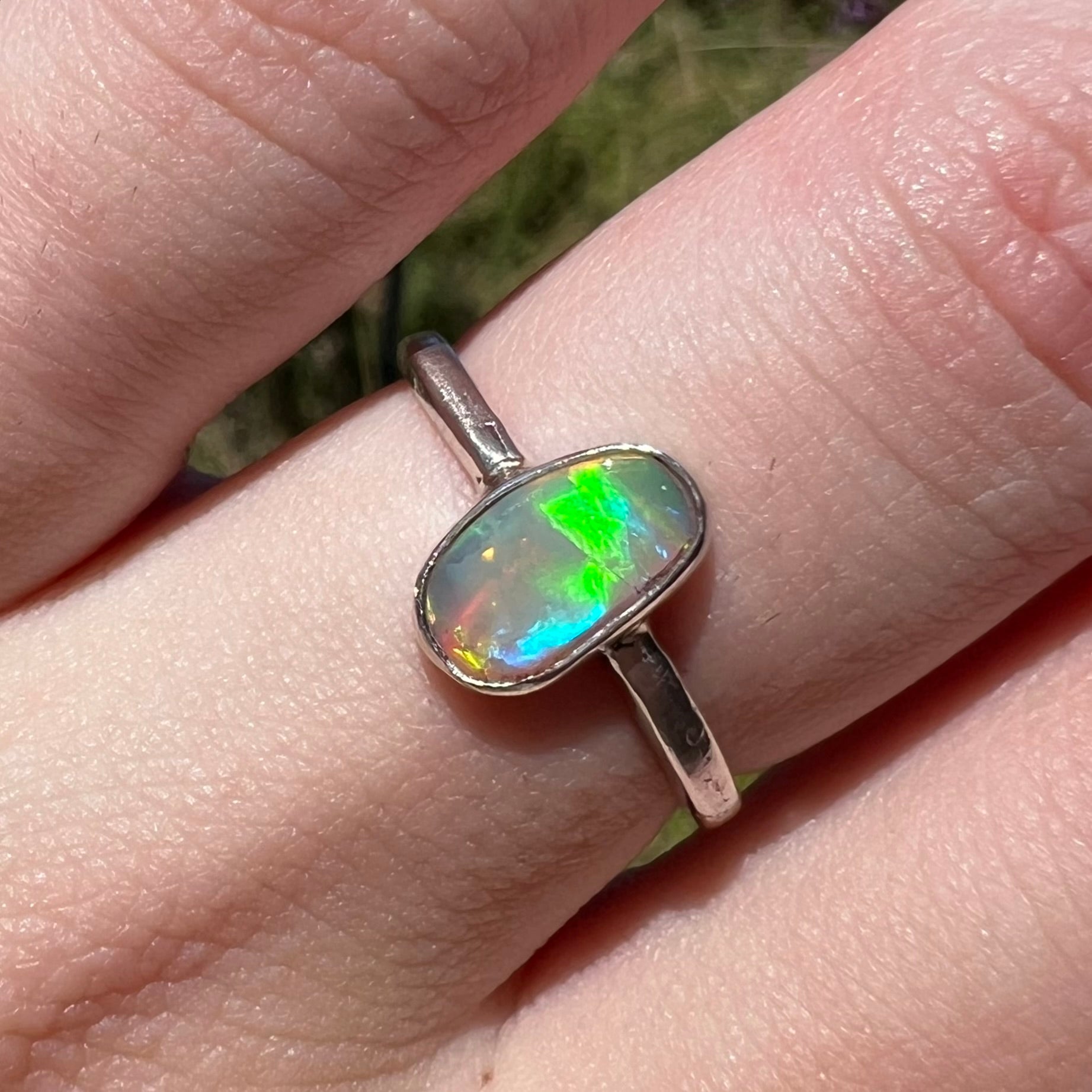 Macro view of an Australian crystal opal women's ring in sterling silver, worn on a finger in sunlight.  The opal has green and blue flashes.