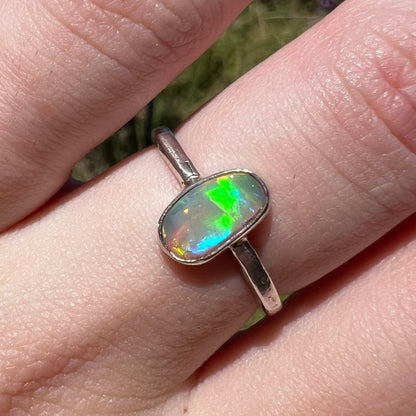 Macro view of an Australian crystal opal women's ring in sterling silver, worn on a finger in sunlight.  The opal has green and blue flashes.