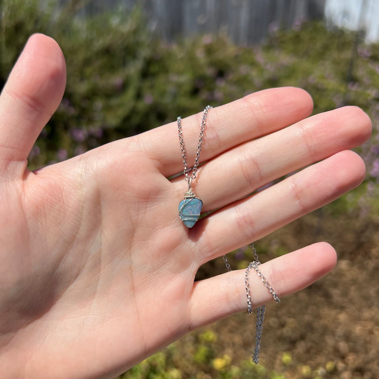 Hand holding an Australian dark opal doublet pendant with a sterling silver wire wrapped design under natural sunlight against blurred outdoor backdrop.