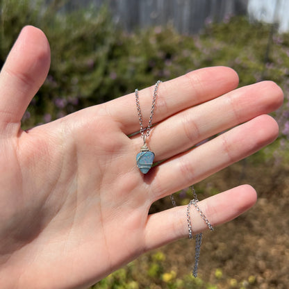 Hand holding an Australian dark opal doublet pendant with a sterling silver wire wrapped design under natural sunlight against blurred outdoor backdrop.